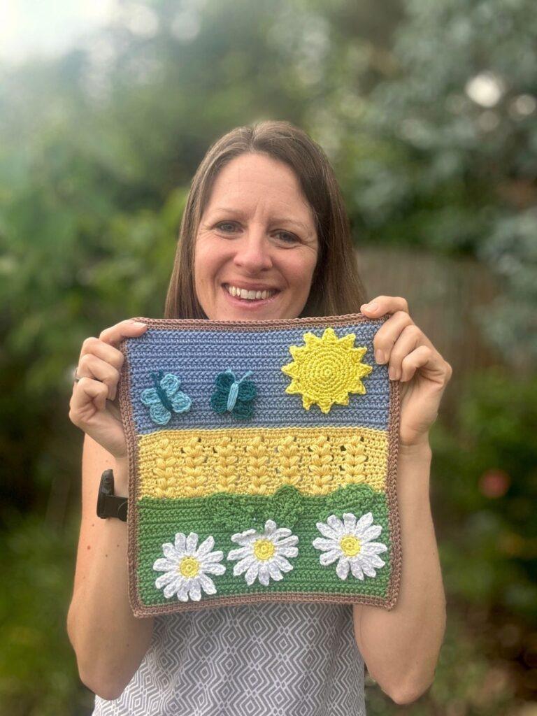 A lady holds up a crochet picture featuring large crochet daisies, a wheat field, butterflies and sun on a crochet background