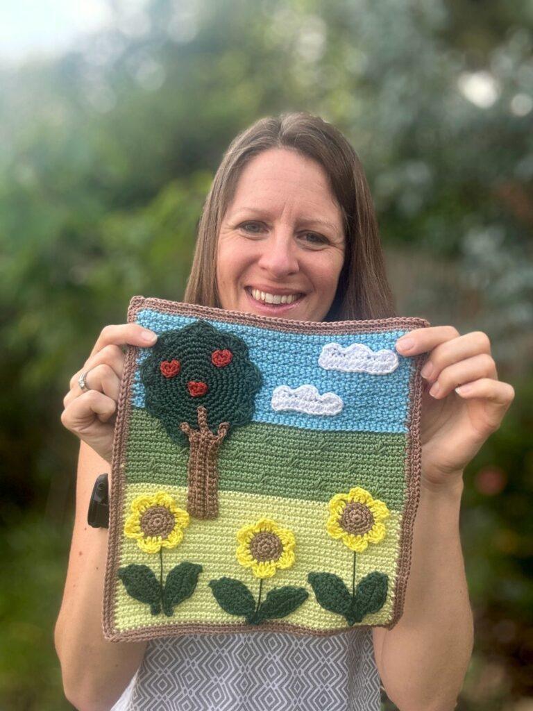 A lady holds up a crochet landscape of a field, tree, clouds and sunflowers