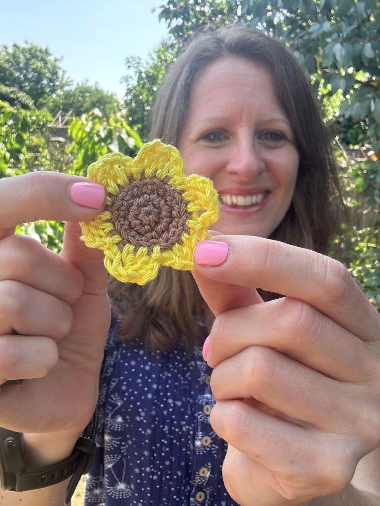 A woman is smiling and holding a yellow and brown crochet sunflower motif out in front of her