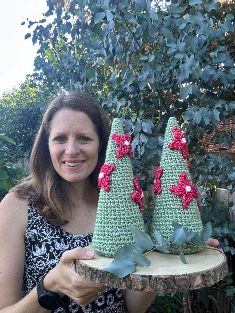 A lady holds a wooden slice with 2 crochet Christmas trees on. The trees have crochet red poinsettia flowers on.