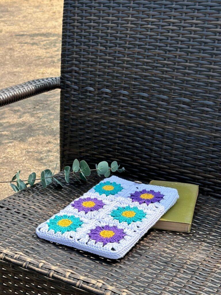 A white, purple and teal floral granny square crochet iPad case lies on a bench with a green book and a sprig of leaves.