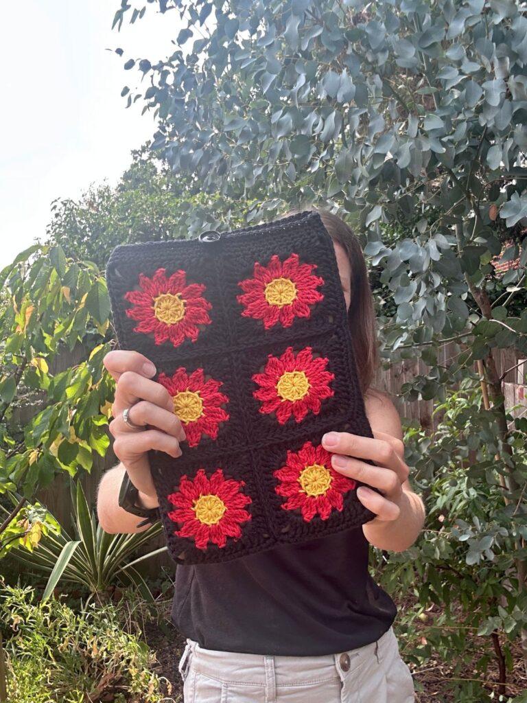 A lady is holding up a crochet floral iPad cover in front of her face, with red and yellow flowers on black background