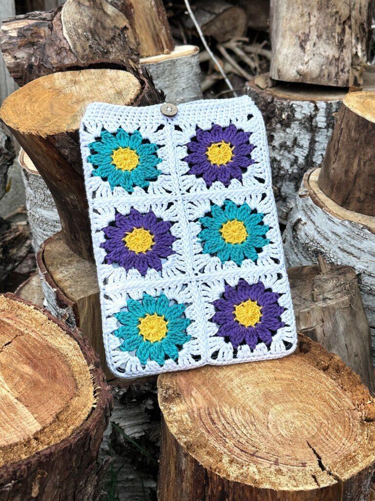 A white iPad case with flower granny squares lays on a pile of wooden logs 