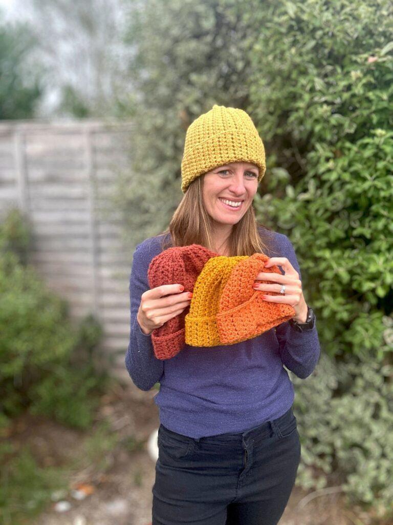 A lady wears a yellow crochet beanie and holds 3 others in different autumnal colours and sizes