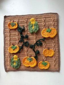 A crocheted picture of a brown field with crochet applique pumpkins, vines and corn on the cobs arranged on it