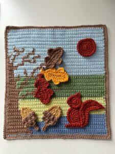 A tapestry crochet picture of a nature scene and a tree, with crochet leaves, acorns and a squirrel on it