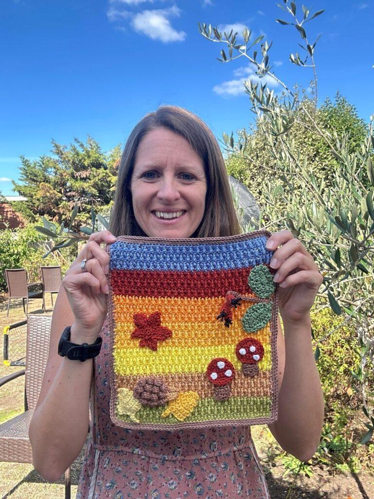 A woman hold a crochet picture featuring rainbow coloured background and crochet mushrooms, hedgehog and leaves