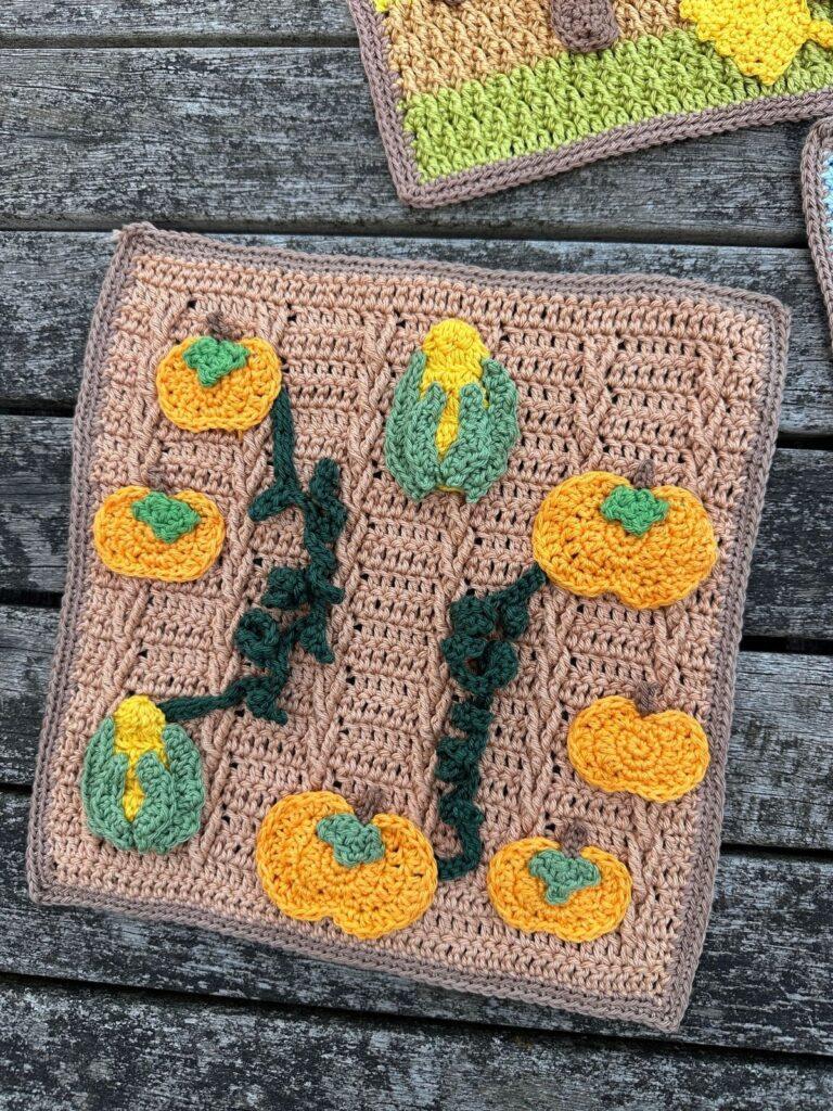 A crocheted picture of a brown field with crochet applique pumpkins, vines and corn on the cobs arranged on it