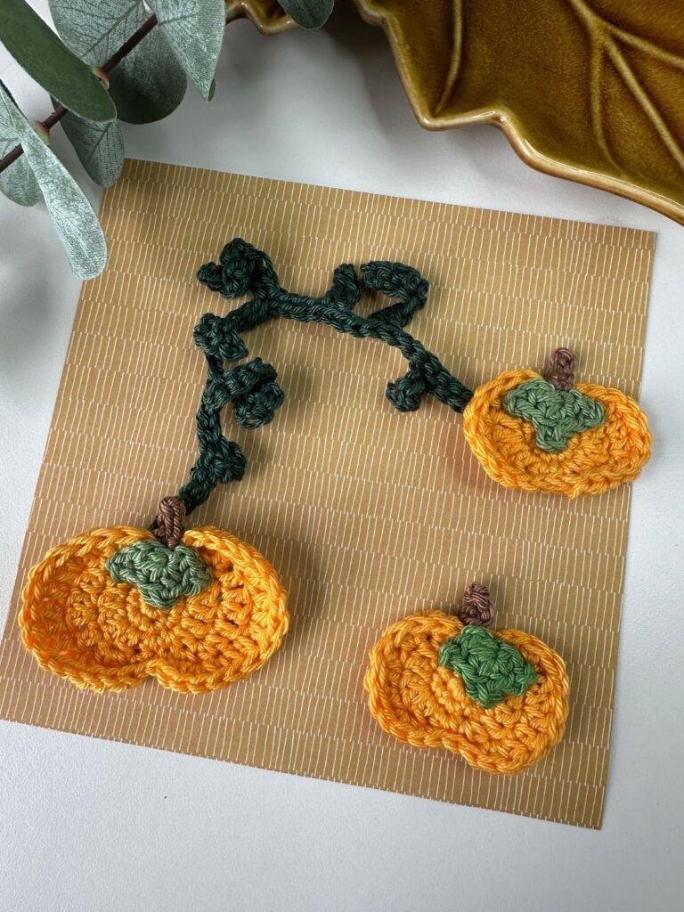 3 cute crochet pumpkin appliqués are laid on a brown piece of paper with twisting crochet vines between them