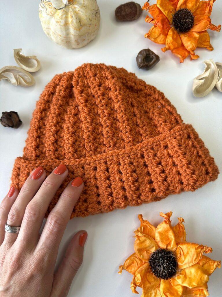 A hand rests on a orange crochet beanie hat with textured ribs and flowers and nuts around the edge
