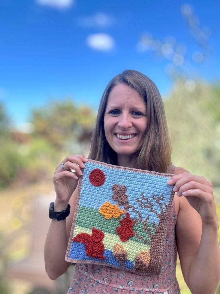 A lady holds up a crochet picture in tapestry crochet of a tree with crochet motifs of leaves, squirrel, acorn and a harvest moon