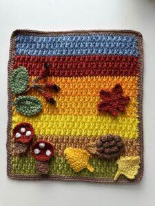 A textured striped crochet landscape background with toadstools, leaves and hedgehog on top