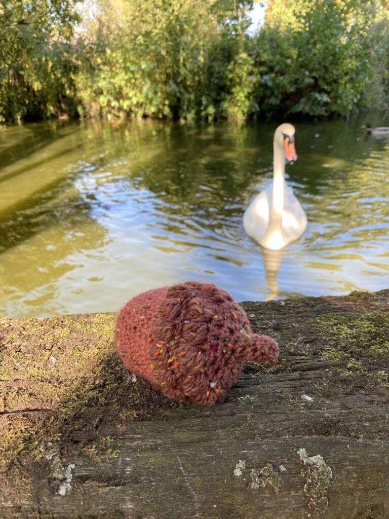 A crochet acorn in brown and red tweed yarn is on a wooden shelf in front of a lake with a swan in the background
