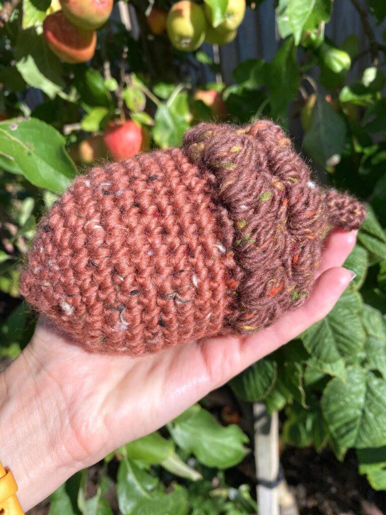 a hand holds a large crochet acorn up against a background of an oak tree