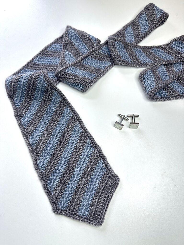 A blue and grey crocheted mens tie on a white surface next to some cuff links