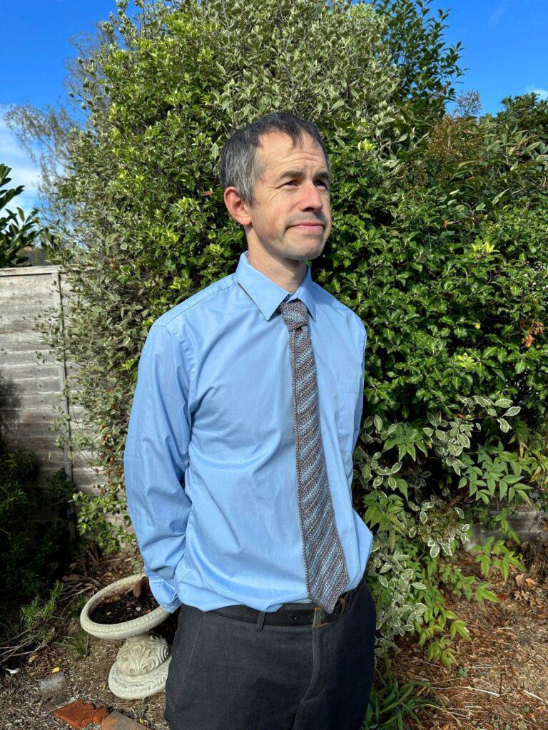 A man stands in a garden wearing a blue shirt and blue and grey striped crochet tie