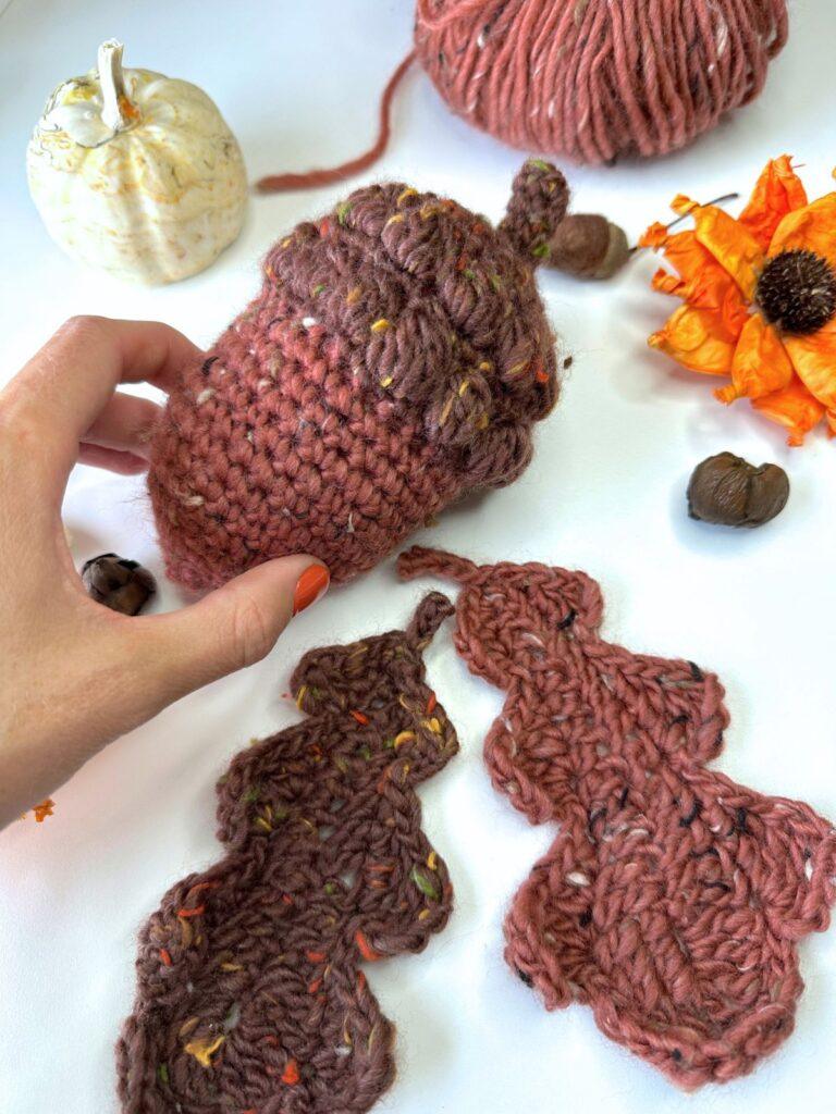 A hand touches a big crochet acorn. Crochet oak leaves and other autumn decorations surround it on a white background