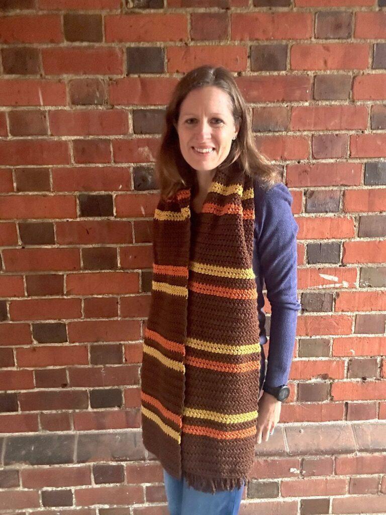 A lady wears a large crochet scarf in brown, yellow and orange stripes