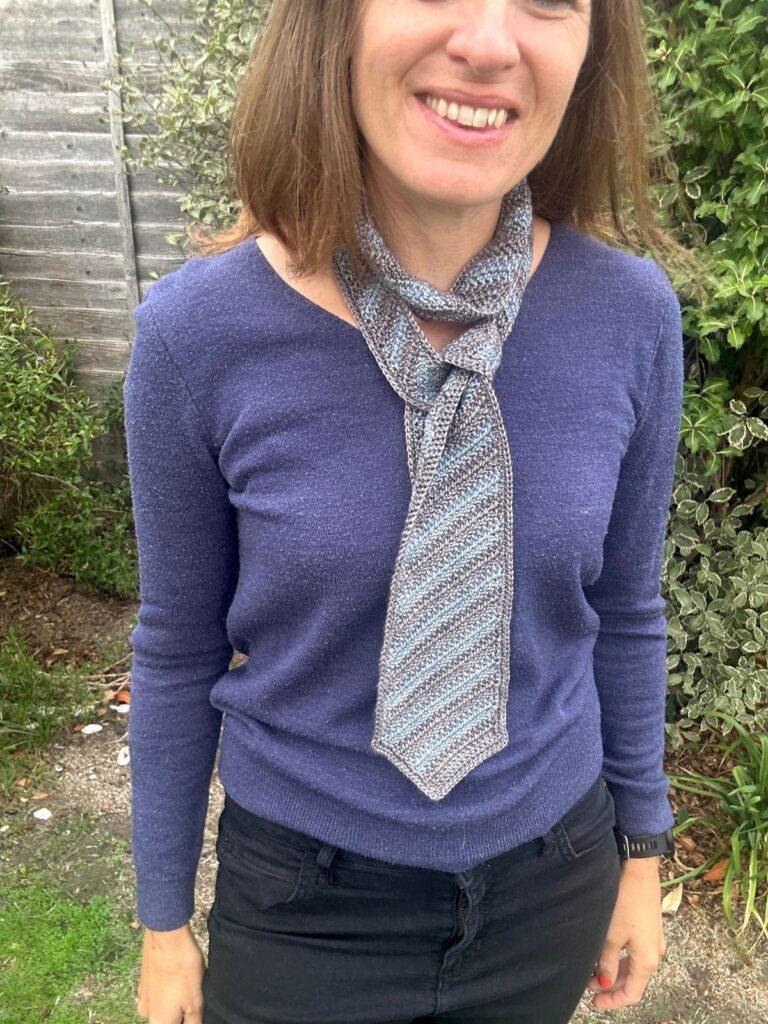 A lady wears a blue sweater and has a crochet tie tie loosely around her neck