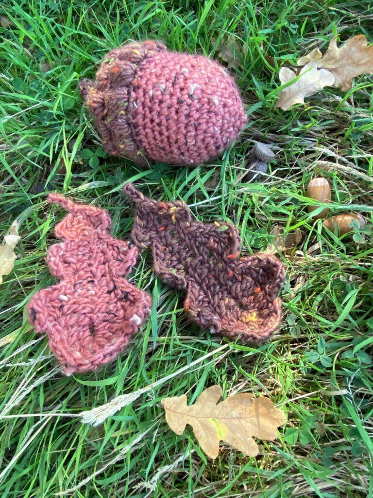 A large crochet acorn and 2 crochet oak leaves lay on the grass with real oak leaves and acorns