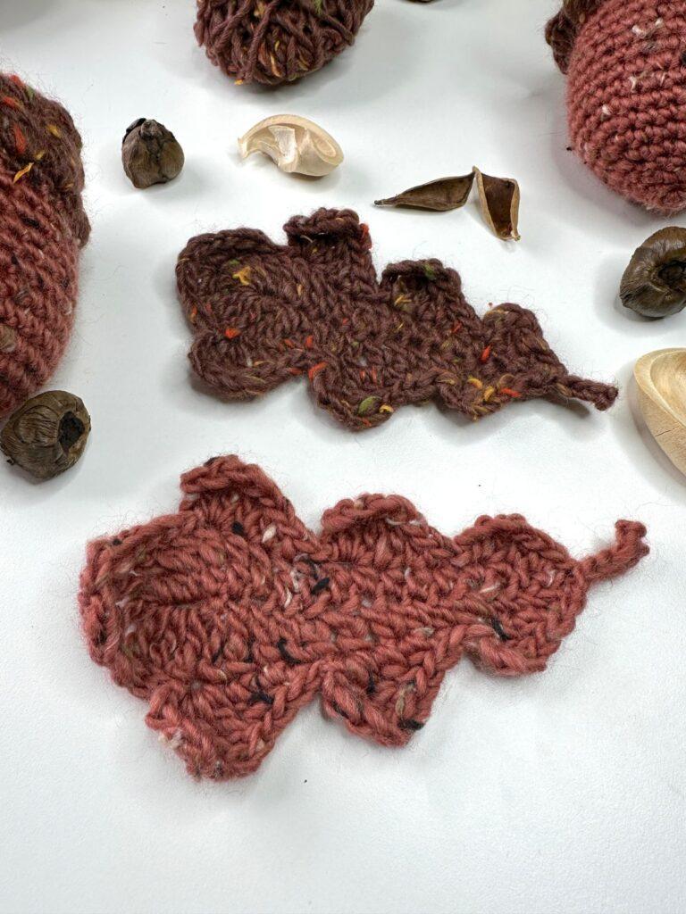 2 crochet oak leaves in tweed yarn lay on a white surface with autumnal nuts and crochet acorns around them