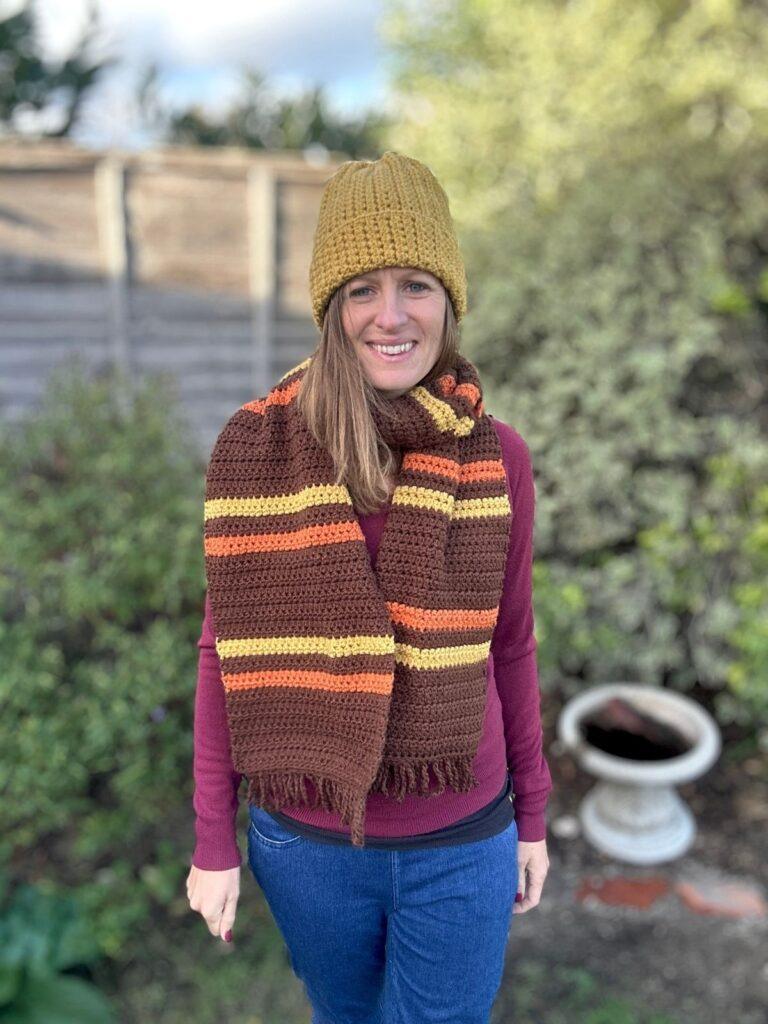 A lady wears a crochet beanie and a brown, yellow and orange striped crochet scarf, which is wrapped round twice and is oversized.