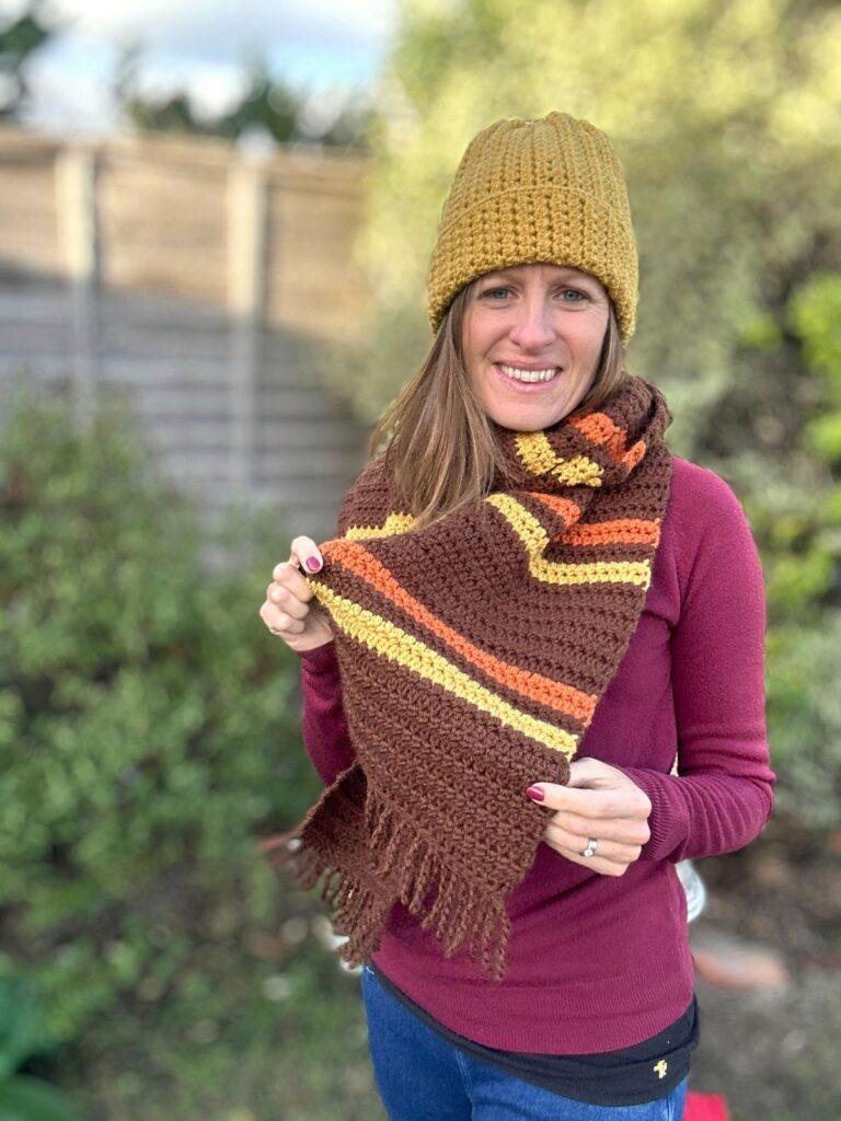 A woman wears a yellow crochet hat and a brown, yellow and orange striped crochet scarf which she holds up to show the twisted fringe.